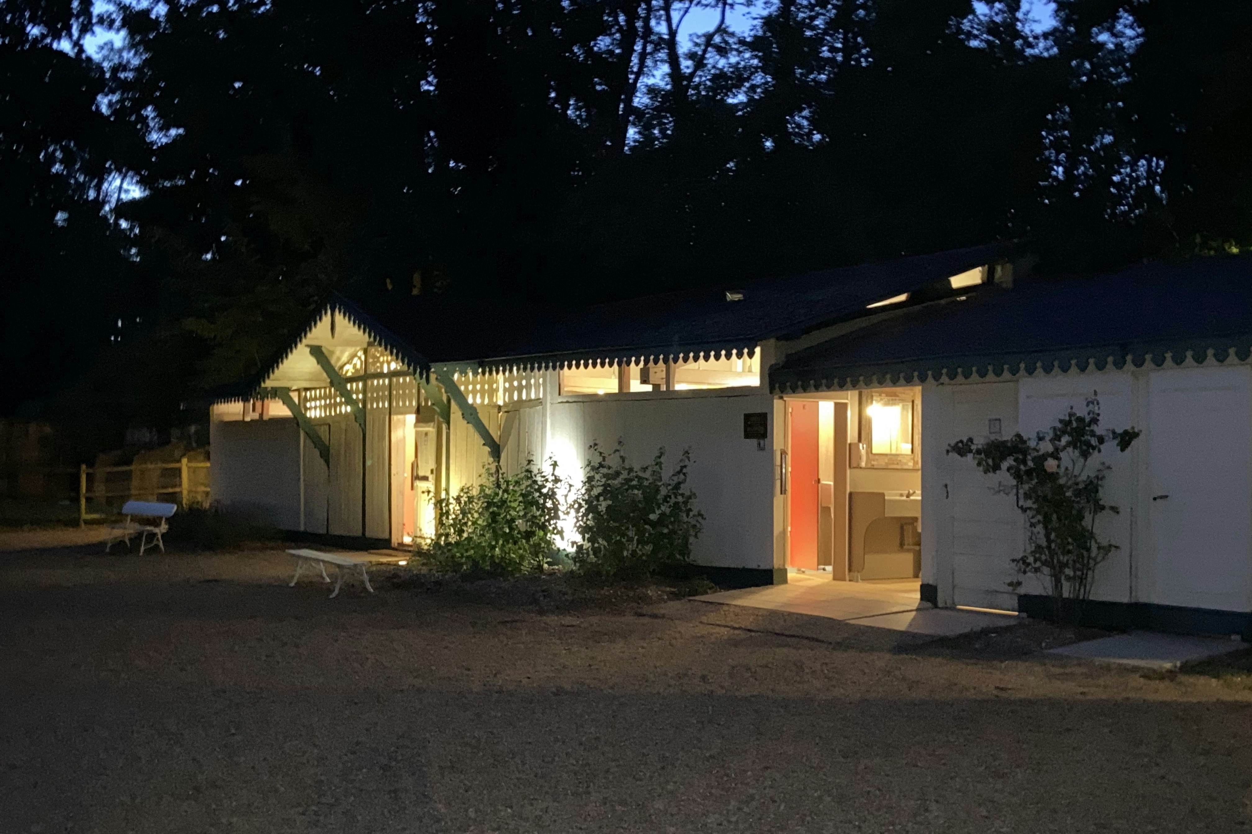 Beleuchtetes Campinghaus mit Terrasse und Eingang, davor Weg und Sitzbank, im Hintergrund Bäume im Dunkeln.