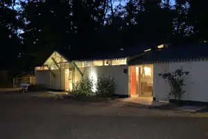 Beleuchtetes Campinghaus mit Terrasse und Eingang, davor Weg und Sitzbank, im Hintergrund Bäume im Dunkeln.