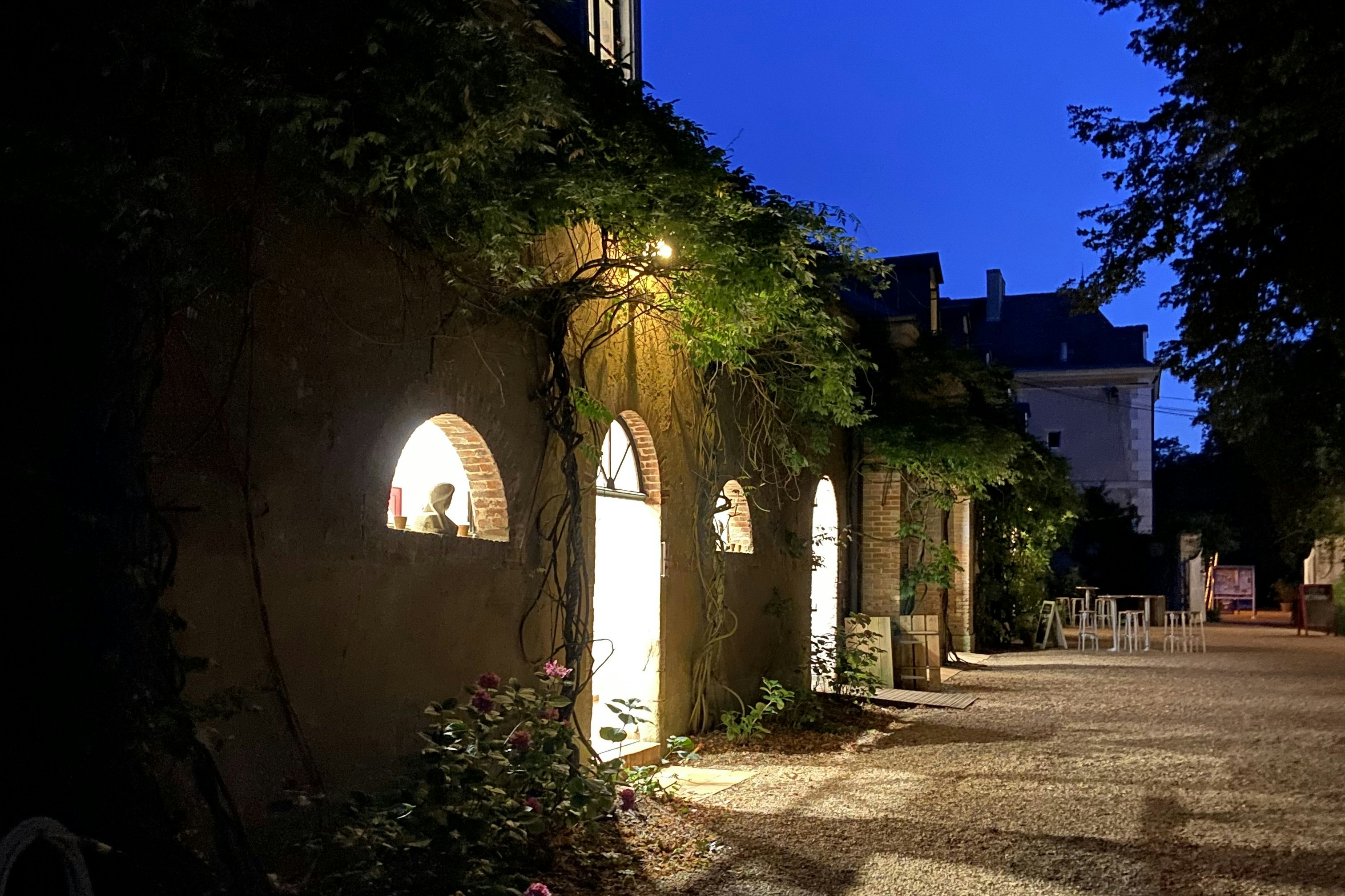 Beleuchteter Steinbogen-Gang am Campingplatz am Abend, mit Sitzgelegenheiten und Bäumen entlang des Weges.