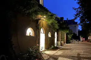 Beleuchteter Steinbogen-Gang am Campingplatz am Abend, mit Sitzgelegenheiten und Bäumen entlang des Weges.