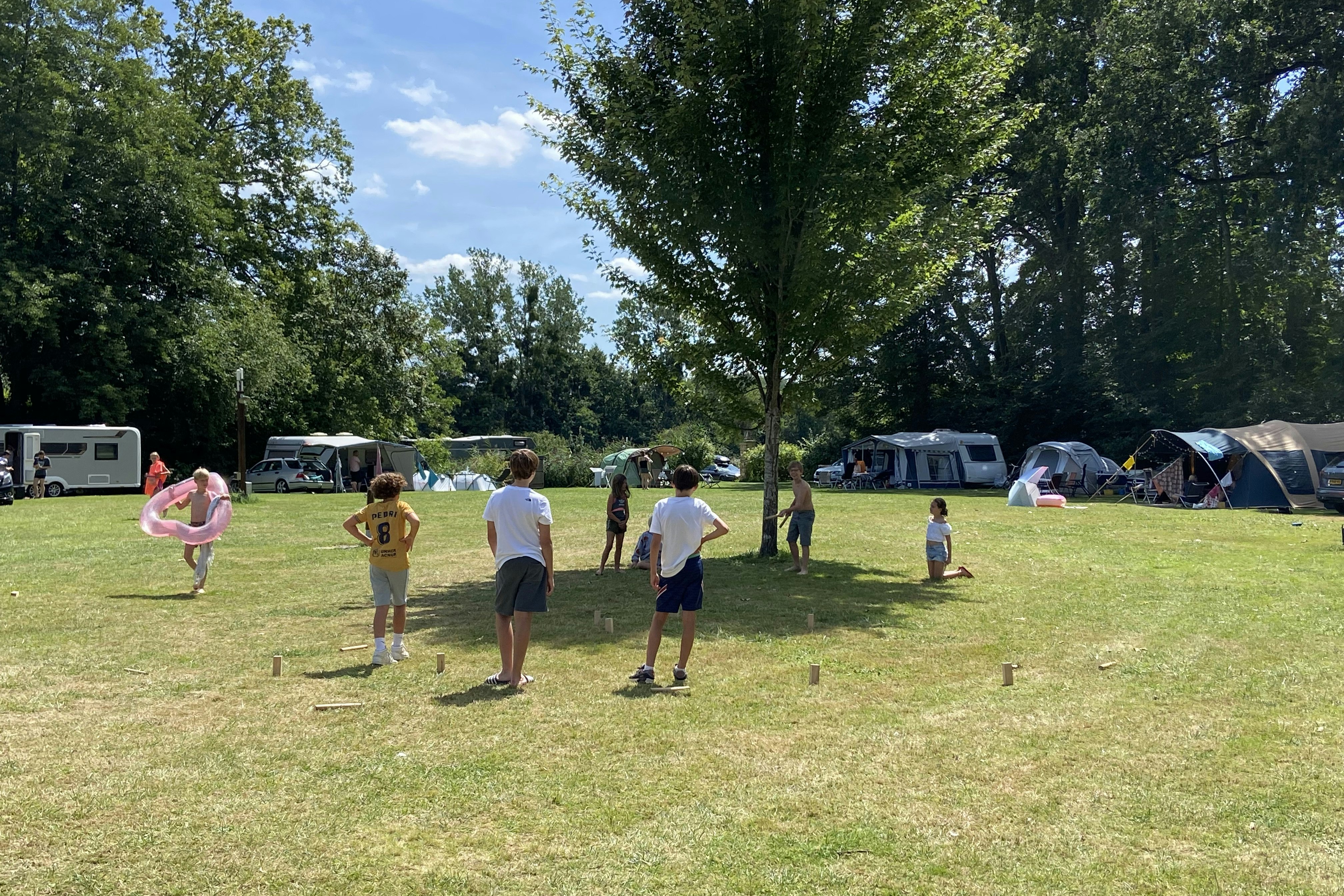 Campingwiese mit Zelten und Wohnwagen, Bäume im Hintergrund und spielende Kinder auf dem Rasen.