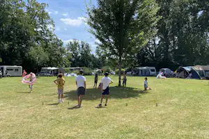 Campingwiese mit Zelten und Wohnwagen, Bäume im Hintergrund und spielende Kinder auf dem Rasen.