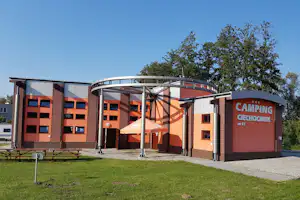 Campinggebäude mit Eingang, orangefarbenen Fassaden und Überdachung; daneben Rasen, Wege und Bänke im Außenbereich.