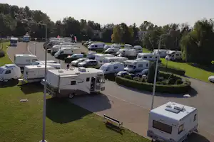 Wohnmobile auf einem Campingplatz, asphaltierter Weg, Heckenbeete und Rasenflächen zwischen Stellplätzen, im Hintergrund weitere Fahrzeuge.