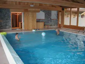 Innenpool mit blauem Wasser, Holzverkleidung und Liegen; zwei Personen planschen im Becken unter einer Holzbalkendecke.
