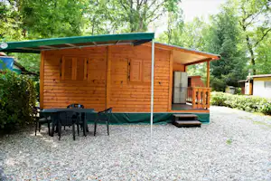 Holzhütte mit überdachter Terrasse, Sitzgruppe und Schiebetür, auf Kiesplatz im Grünen zwischen Bäumen.