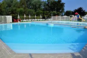 Großes blaues Schwimmbecken auf dem Campingplatz, mit Liegen, Sonnenschirmen und Zaun im Hintergrund.