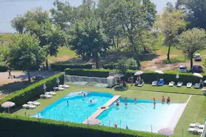 Großes Camping-Schwimmbecken mit Liegen und Sonnenschirmen, umgeben von Rasen, Bäumen und einem gepflegten Gartenbereich.