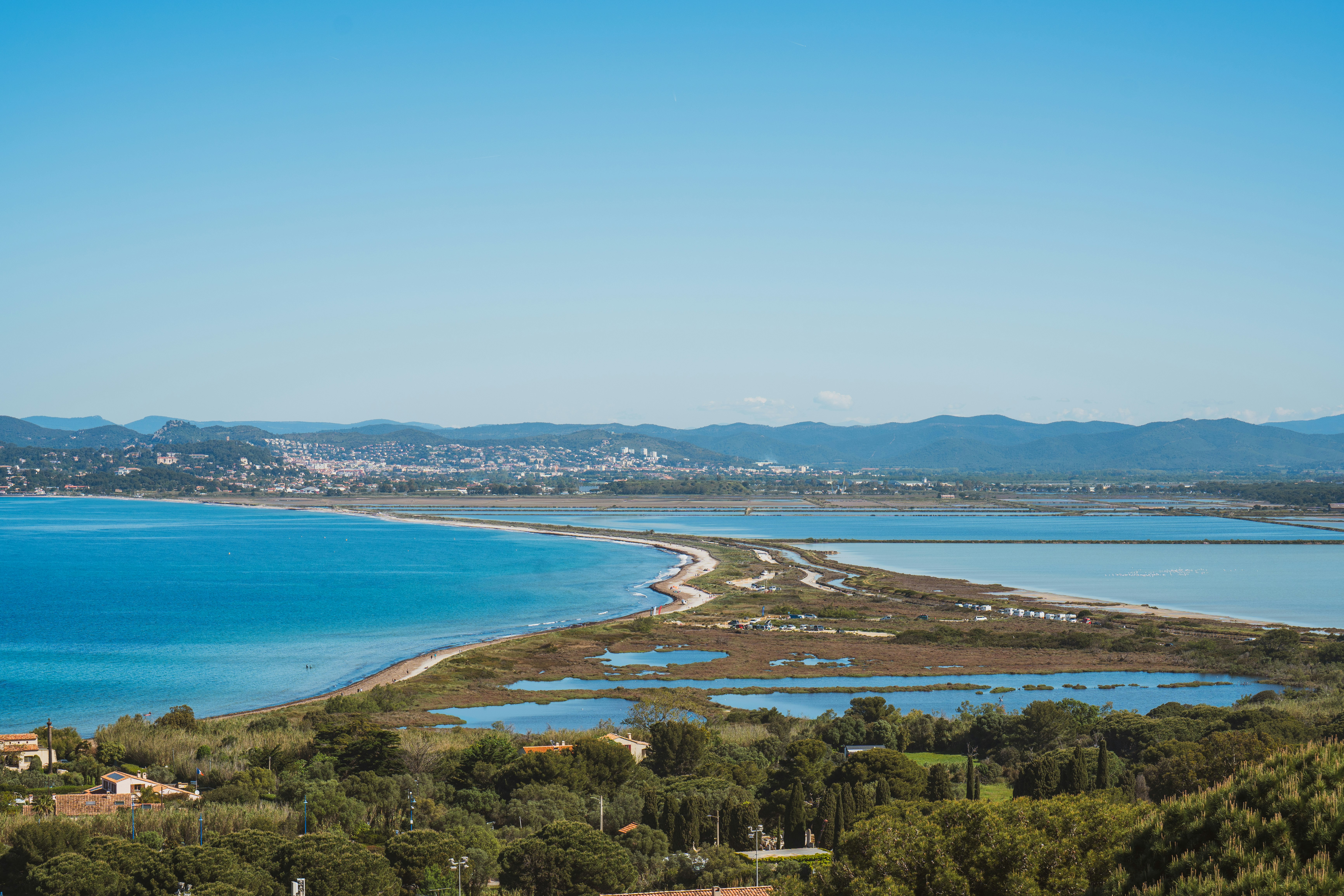 Blick vom Campingplatz auf eine Bucht mit Sandstrand, Lagunenwasser und Küstenlinie, im Hintergrund Berge und Häuser.