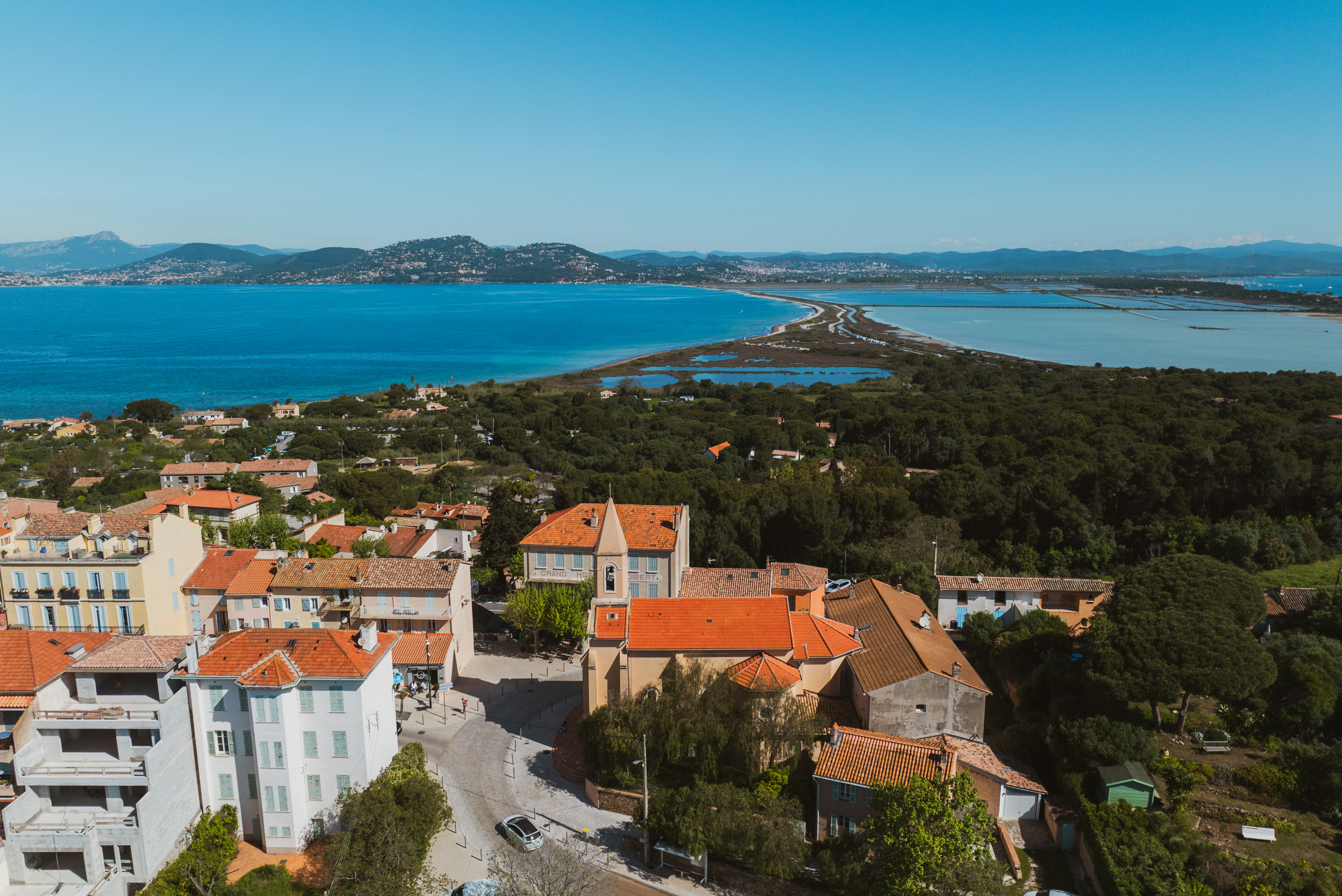 Blick von oben auf ein Küstendorf mit Häusern und roten Dächern, Straße und Meerbucht im Hintergrund.