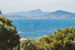 Blick vom Campingplatz auf das Meer mit Bergen im Hintergrund, umgeben von Bäumen und Häusern mit roten Ziegeldächern.