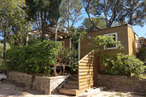 Holzbungalow mit Stufen und kleiner Terrasse, umgeben von Hecken und Bäumen im Campingbereich.