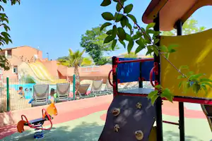 Spielbereich mit Rutsche und Kletterwand auf dem Campingplatz, im Hintergrund Pool, Liegen und Zäune.