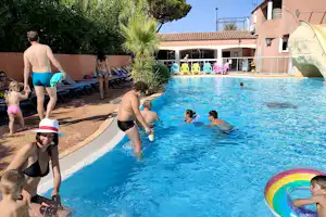 Familie im Außenpool, Kinder mit Schwimmreifen, Sonnenschirme und Liegen am Beckenrand, Gebäude mit Terrasse im Hintergrund.