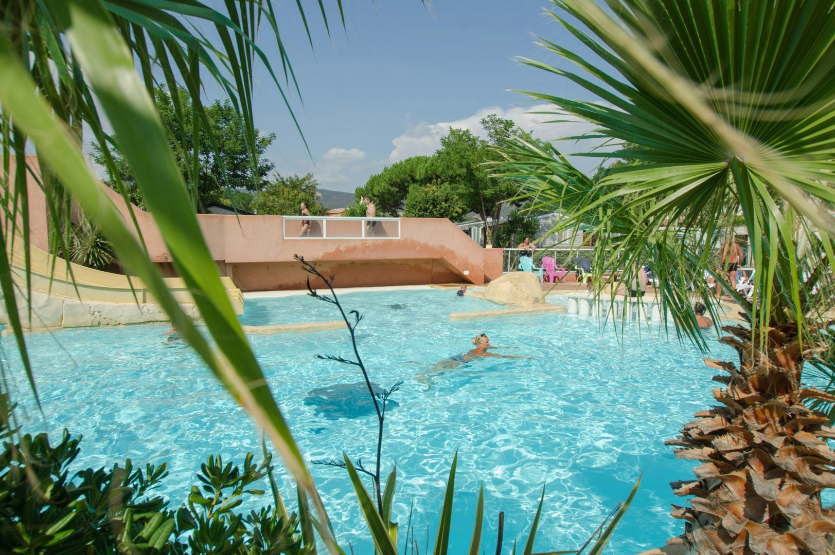 Blauer Camping-Pool mit Palmen im Vordergrund und Liegen am Beckenrand, dahinter ein helles Gebäude.