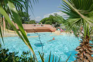 Blauer Camping-Pool mit Palmen im Vordergrund und Liegen am Beckenrand, dahinter ein helles Gebäude.
