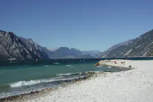 Schotterstrand am See mit türkisfarbenem Wasser, Booten und Bergen im Hintergrund unter klarem Himmel.