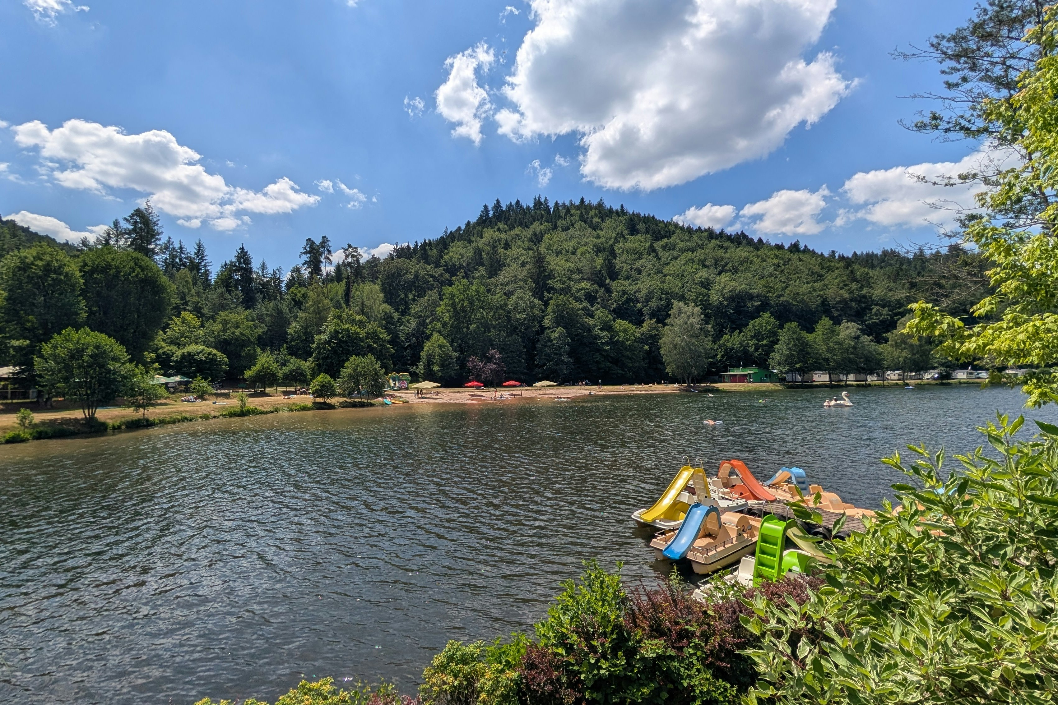 Ruhiger See mit gelbem Schlauchboot am Ufer, umgeben von Bäumen und grünen Hügeln unter bewölktem Himmel.