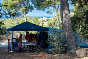 Camping unter einem blauen Zelt mit Tisch und Stühlen, große Bäume und Blick auf das Meer in der Ferne.