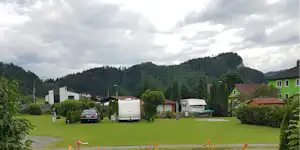 Wohnwagen-Stellplatz auf grünem Campinggelände mit Hecken, Gartenmöbeln und mehreren Fahrzeugen vor bewaldeten Bergen.