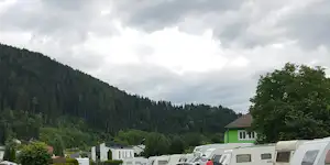 Reihe Wohnwagen auf dem Campingplatz, im Hintergrund bewaldeter Hang und ein grünes Haus mit Dachziegeln.