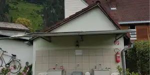 Außenwand eines Campinggebäudes mit Dachvorsprung, Fliesenwand, Feuerloescher und Fahrrad am Eingang, im Hintergrund grüne Berge.