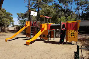 Gelbes Spielgerät mit Rutschen und Kletterelementen auf einem Sandplatz, umgeben von Bäumen und Zaun.