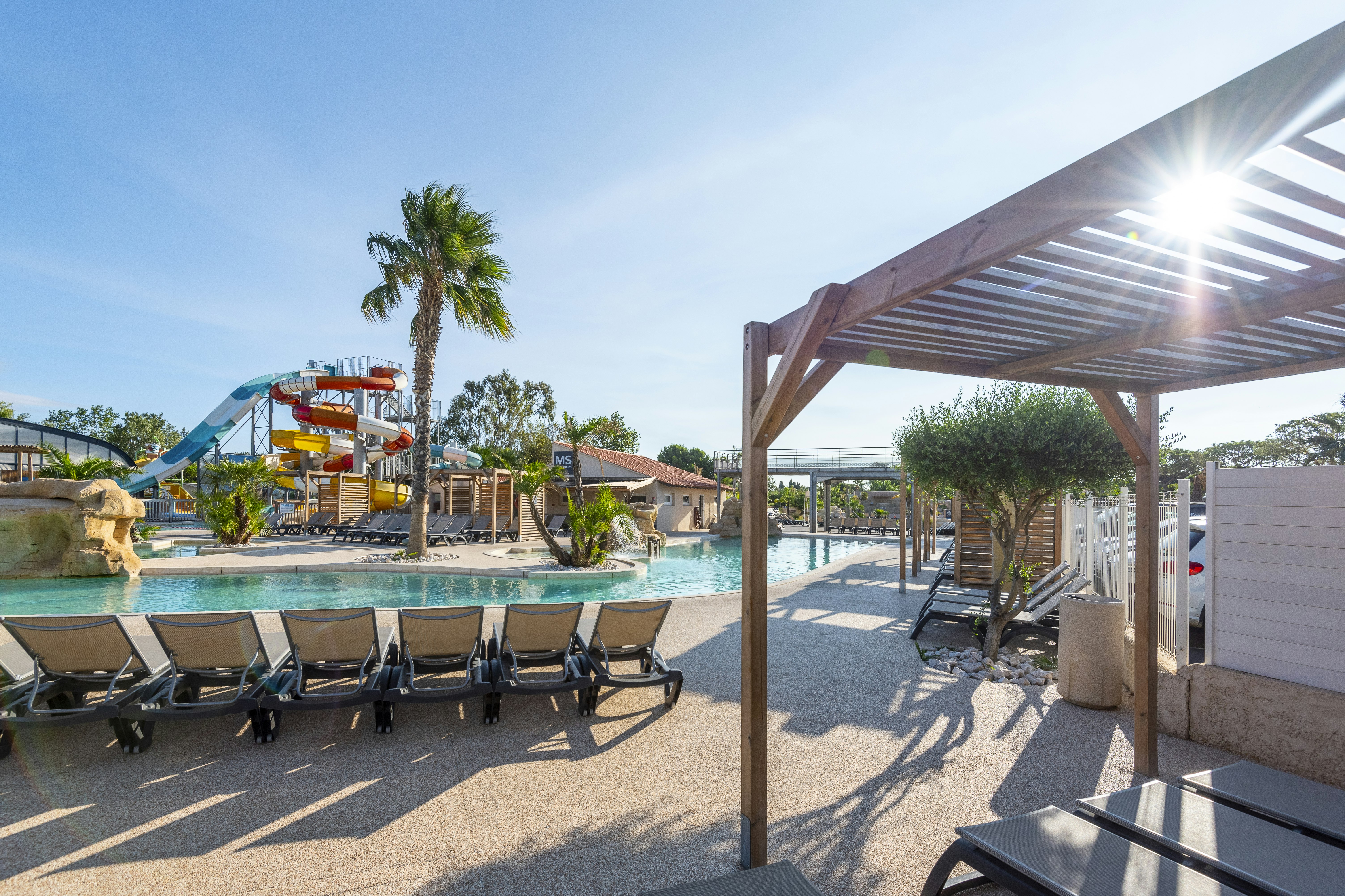 Poolbereich mit Sonnenliegen und Sonnenschirmen, Holzpergola am Rand, im Hintergrund Spielplatz und Palmen vor Campinganlage.