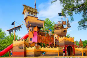 Holz-Spielburg mit Rutsche und Kletterwänden auf dem Campingplatz, mit Bäumen im Hintergrund und mehreren Eingängen.