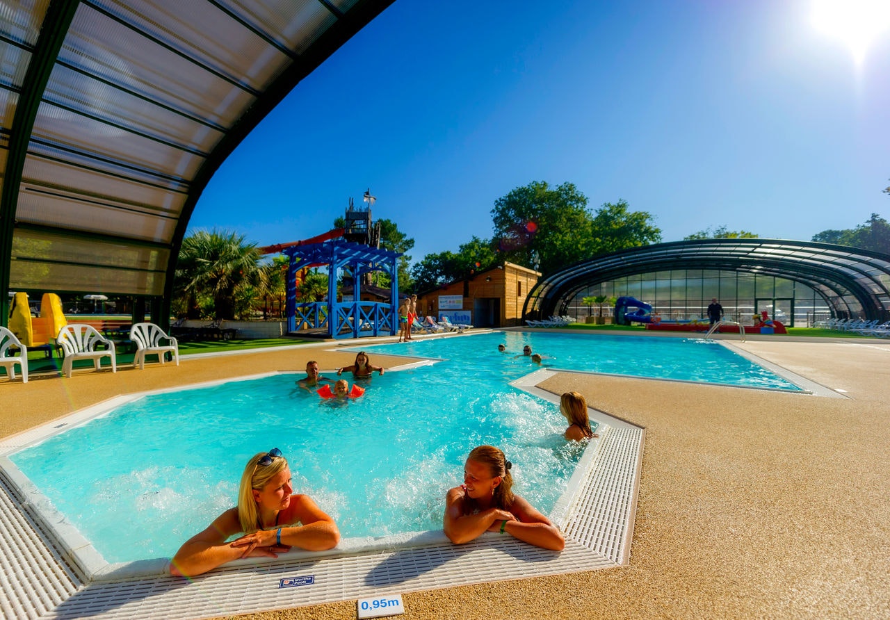 Kinder und Erwachsene im Schwimmbecken eines Campingplatzes, Sonnenschirme und Liegen am Pool, Glasüberdachung und Gebäude im Hintergrund.