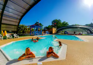 Kinder und Erwachsene im Schwimmbecken eines Campingplatzes, Sonnenschirme und Liegen am Pool, Glasüberdachung und Gebäude im Hintergrund.