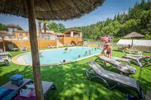 Camping-Pool mit türkisfarbenem Wasser, Liegen auf dem Rasen und Sonnenschirm, im Hintergrund Zelte und Gebäude.