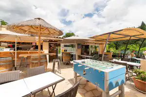 Terrasse am Campingplatz mit Tischkicker, Holzbänken und Sonnenschirmen, daneben eine Bar mit Theke und Sitzgelegenheiten.