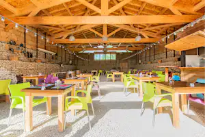 Überdachter Grillbereich mit vielen bunten Tischen und Stühlen, Steinmauer, Holzdecke und Lichterkette im Innenraum.