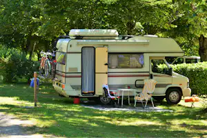 Wohnmobil auf einem Campingplatz im Grünen, mit Markise, Campingstühlen und Tisch am Stellplatz zwischen Bäumen.
