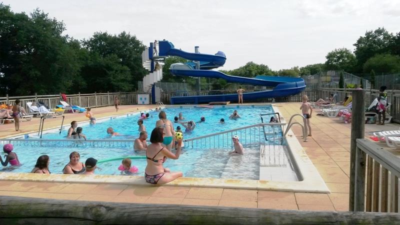 Schwimmbecken auf dem Campingplatz mit blauen Wasserrutschen, Liegen am Beckenrand und mehreren Personen im Wasser.