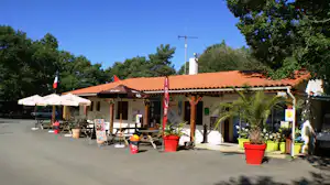 Überdachter Eingangsbereich am Campingplatz mit Sitzgelegenheiten, Sonnenschirmen, Töpfen und Bäumen im Hintergrund.