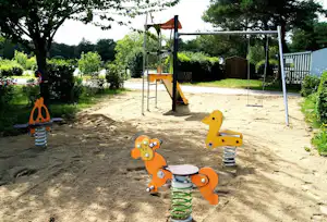 Spielplatz im Campinggarten mit Federwipp-Tieren, Sandboden und Schaukel unter Bäumen.