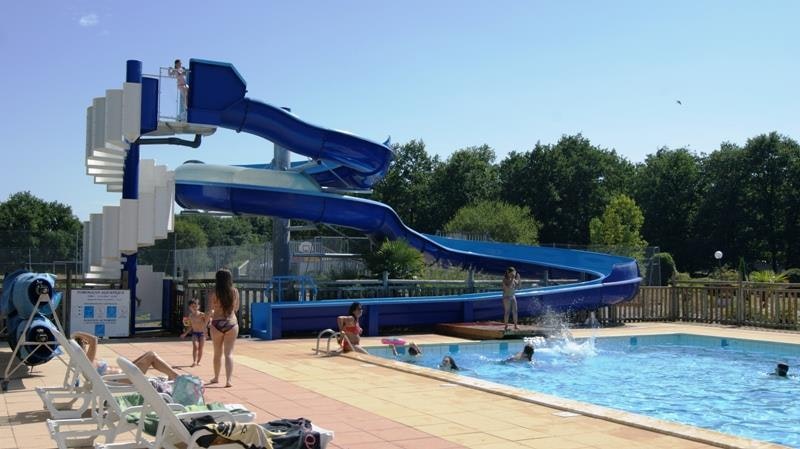 Blaues Schwimmbad mit mehreren Wasserrutschen, Liegen am Beckenrand und Zaun vor Bäumen im Hintergrund.