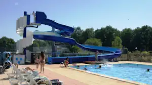 Blaues Schwimmbad mit mehreren Wasserrutschen, Liegen am Beckenrand und Zaun vor Bäumen im Hintergrund.