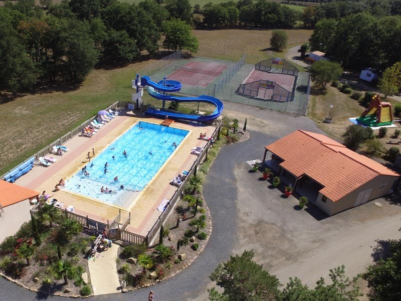 Luftaufnahme eines Campingplatzes mit großem Pool, Liegen am Beckenrand, Wegen und einem Gebäude mit Terrasse neben Bäumen.