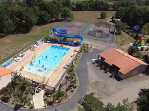 Luftaufnahme eines Campingplatzes mit großem Pool, Liegen am Beckenrand, Wegen und einem Gebäude mit Terrasse neben Bäumen.