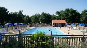 Schwimmbecken auf einem Campingplatz, blaue Sonnenschirme, Liegen und Zäune, im Hintergrund Bäume und ein Gebäude mit rotem Dach.