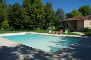 Camping-Pool mit türkisfarbenem Wasser, Sonnenliegen und Zaun, im Hintergrund Bäume und ein Gebäude mit Dachziegeln.