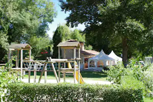 Spielplatz mit Holzschaukel und Rutsche, Zelte im Hintergrund und Bäume rund um den Campingplatz.