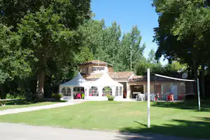 Weißes Zelt vor einem Campinghaus mit Terrasse, Gartenrasen und vielen Bäumen im Hintergrund.