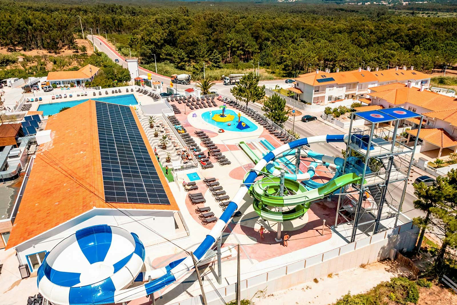 Großer Campingbereich mit beheiztem Pool, Sonnenterrassen, Liegen und orangefarbenen Gebäuden, umgeben von Bäumen und Hügeln.