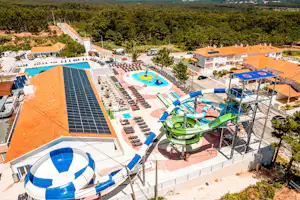 Großer Campingbereich mit beheiztem Pool, Sonnenterrassen, Liegen und orangefarbenen Gebäuden, umgeben von Bäumen und Hügeln.