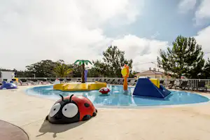 Kinderfreundlicher Pool mit Spielzeug-Ente und Schwimmspielzeug auf dem Betonrand, daneben Liegen und Sonnenschirme.