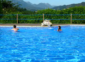 Blauer Campingpool mit zwei Personen im Wasser, im Hintergrund ein Wasserstrahl und Berge.